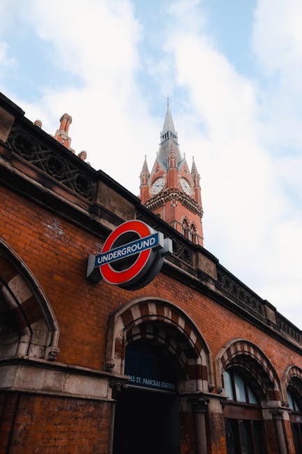 A close-up view of a brick building with arched windows and a prominent London Underground sign attached to its facade. Behind the building, part of a historic clock tower with a pointed spire is visi