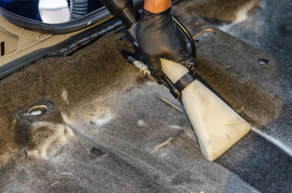 A person is using a yellow industrial vacuum cleaner with a black hose and a wide cleaning head to deep clean a patterned carpet in a residential setting. The carpet features a floral design in subdue